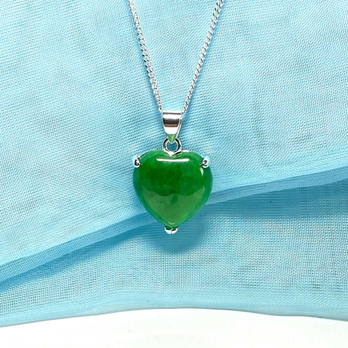 Green heart-shaped pendant on a silver chain against a light blue fabric background