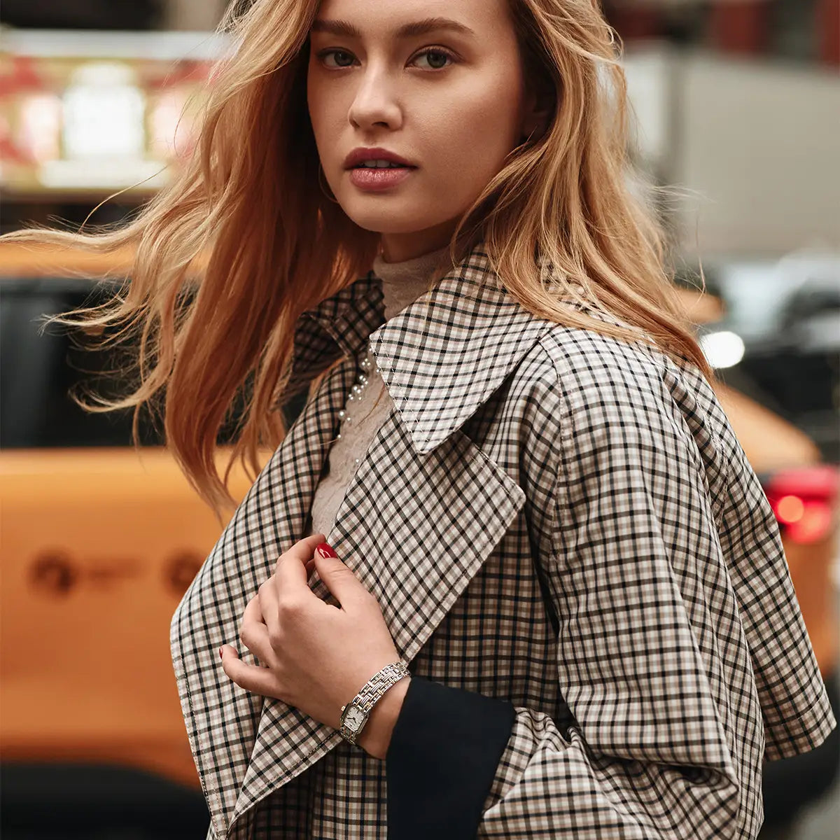 A woman wearing a beige and black checkered coat over a black top, photographed in an outdoor urban setting with a blurred orange taxi in the background.