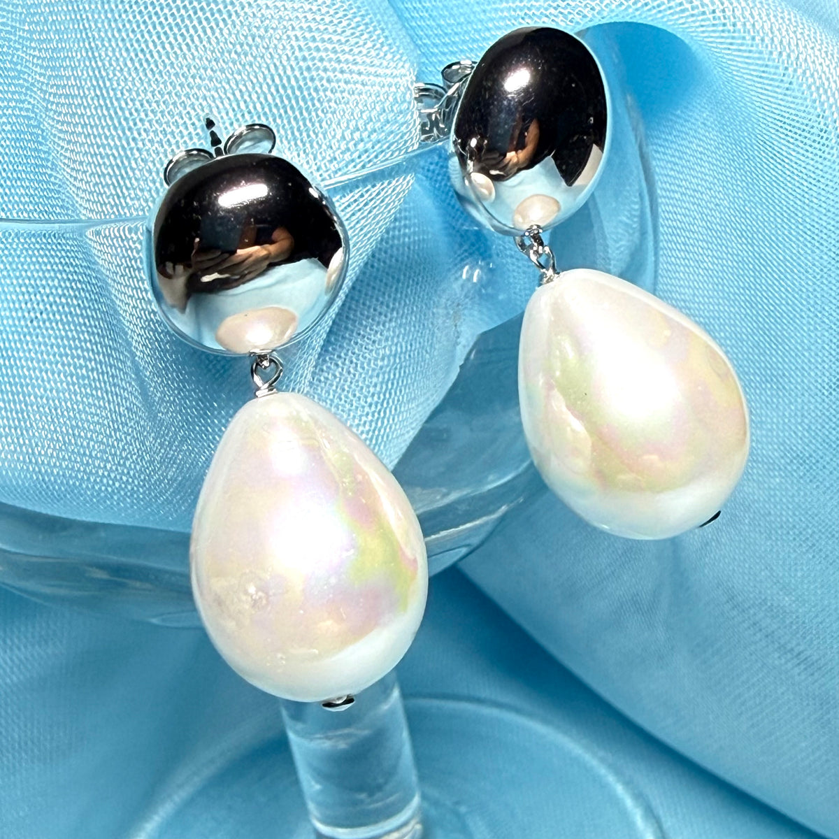 A pair of silver drop earrings with large, lustrous white baroque pearl teardrops hanging from polished silver oval studs, displayed on a clear glass stem against a light blue fabric background.