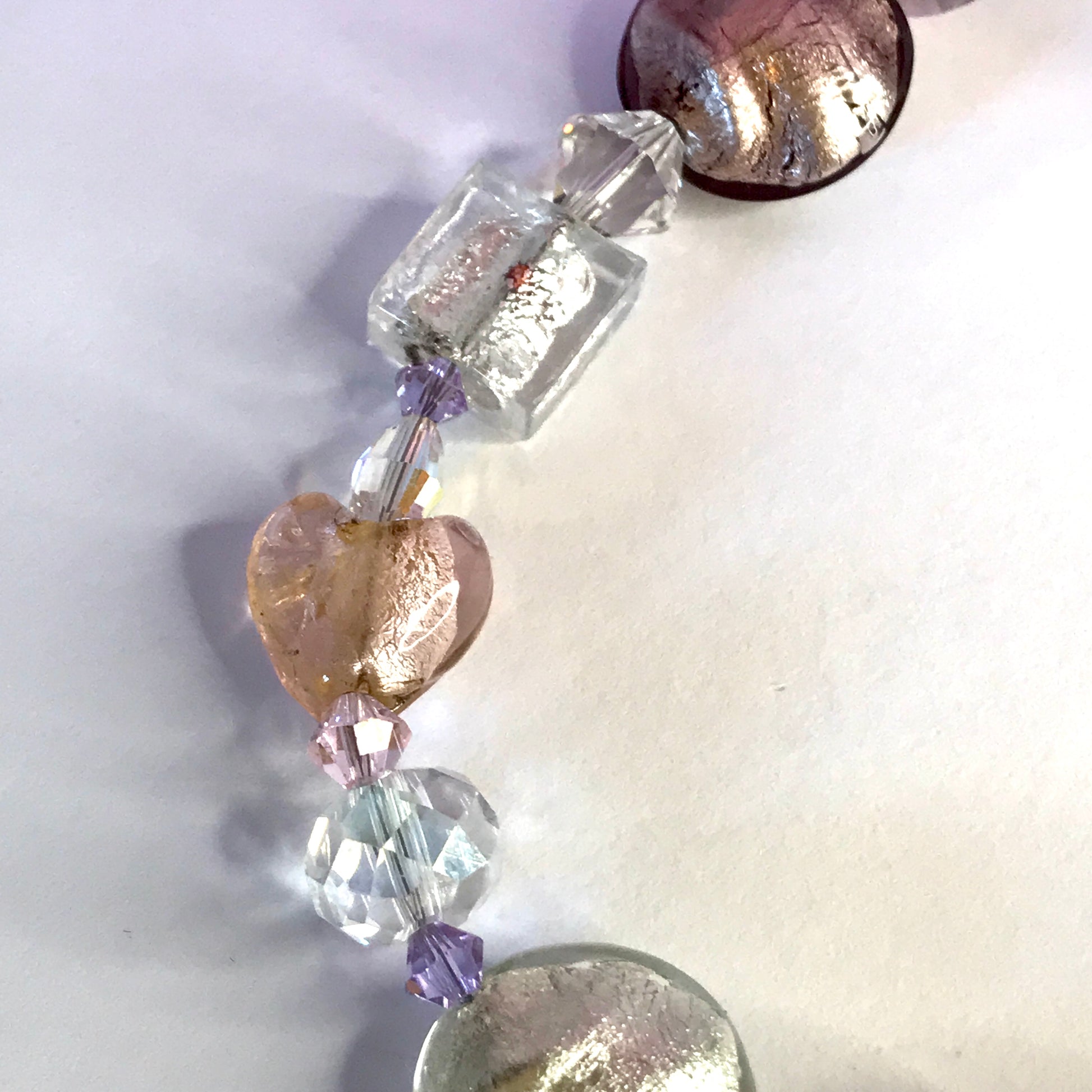 A close-up of a beaded strand featuring clear faceted beads, a textured rose-gold heart bead, a light pink faceted bead, a small lavender purple bead, and a large round textured metallic bead at the bottom.
