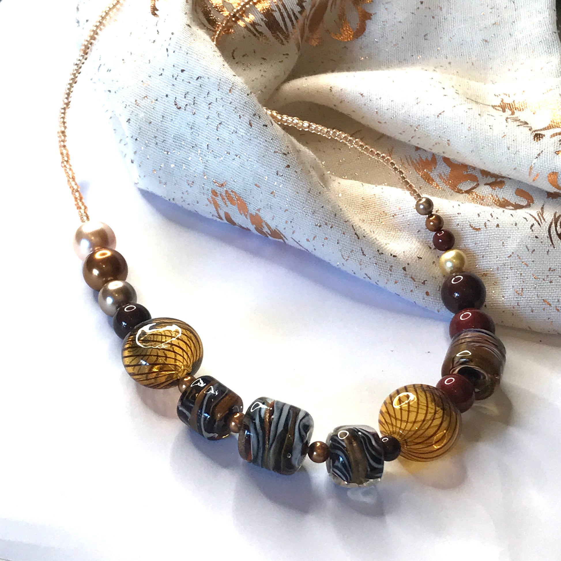 A beaded necklace with a gold-tone chain, featuring a mix of textured glass beads in brown, black, white, and amber tones, draped over a cream-colored patterned fabric.