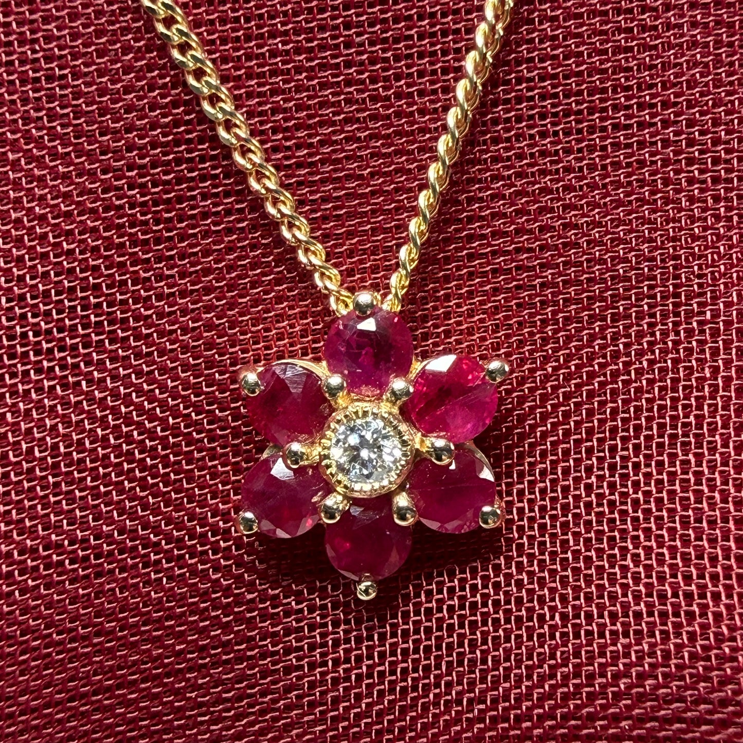 Gold necklace with a flower-shaped pendant featuring red gemstones and a central diamond on a textured red background.