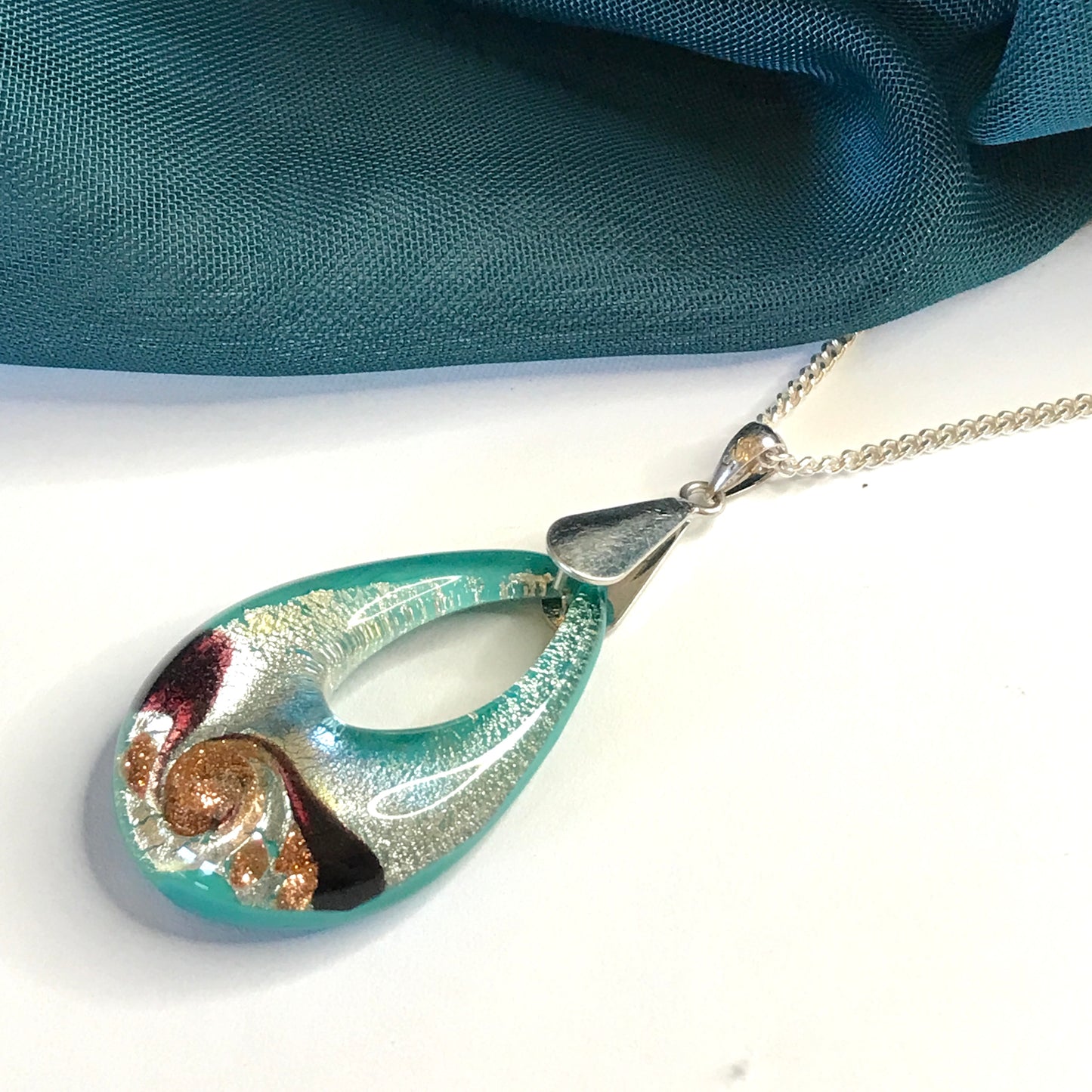 Real Murano glass necklace teal and silver pendant on a white surface with a blue fabric background
