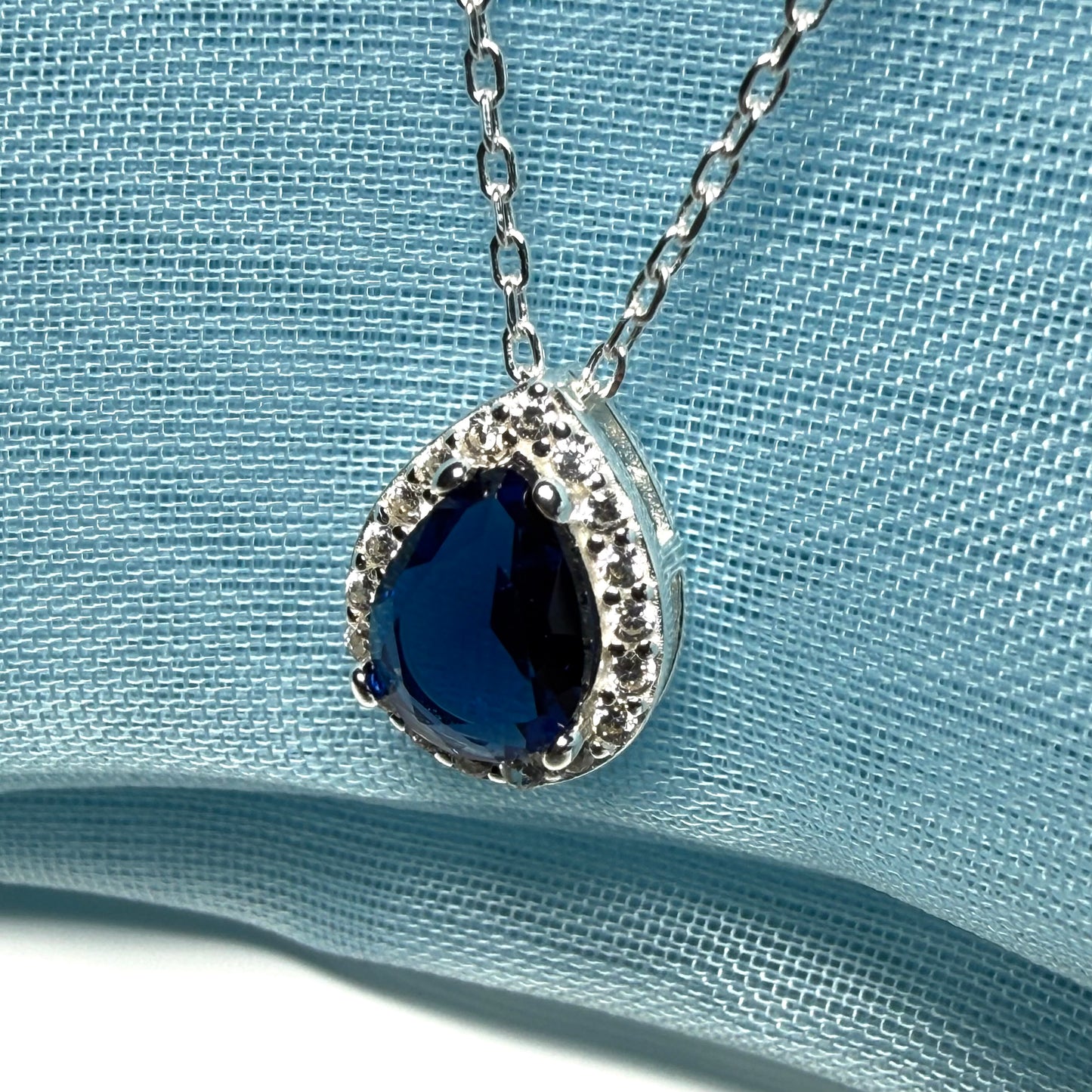 Silver necklace with a blue teardrop pendant on a light blue fabric background