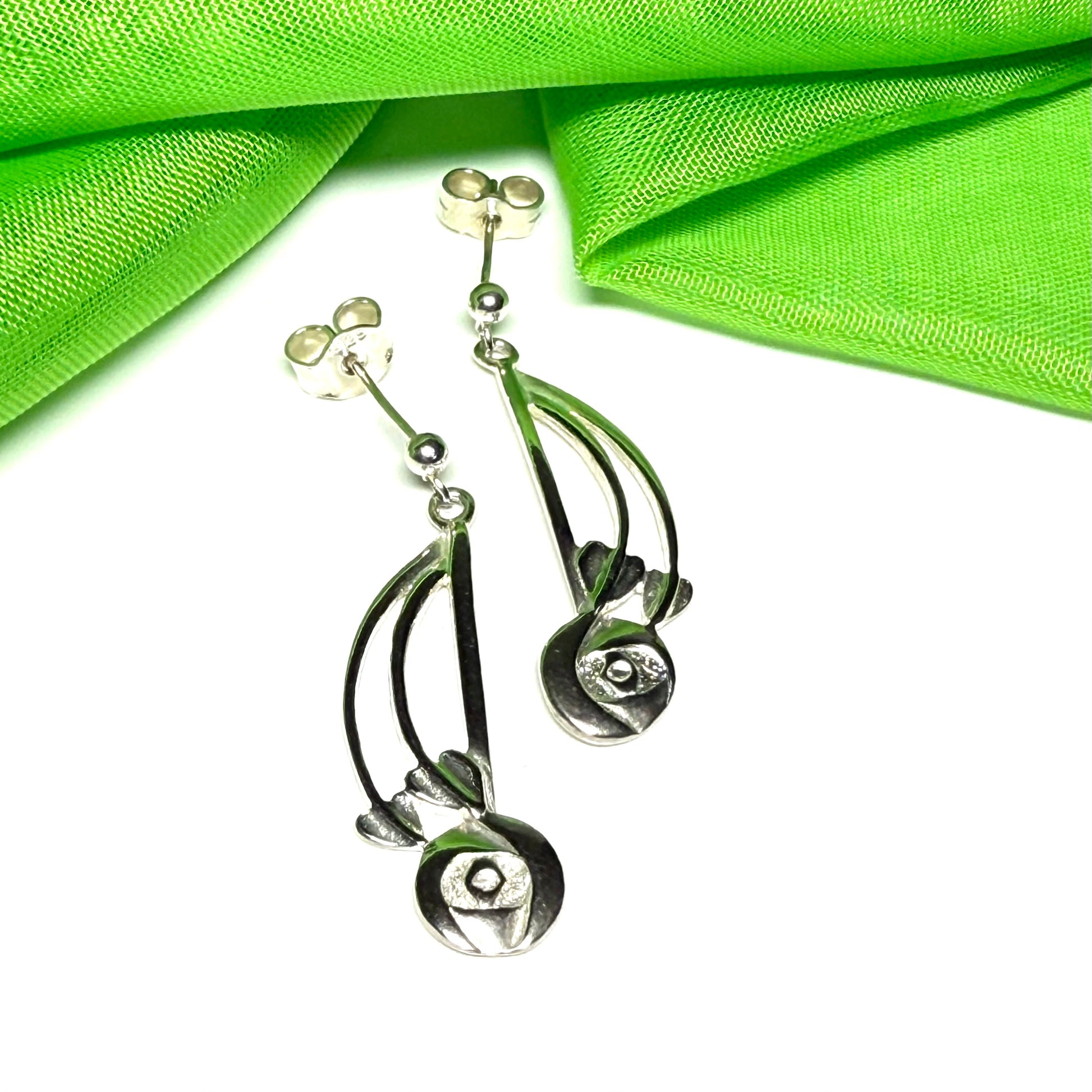 A pair of silver drop earrings with curved leaf-like metal elements and oval dangling charms, displayed on a white background with bright green fabric in the upper corners.