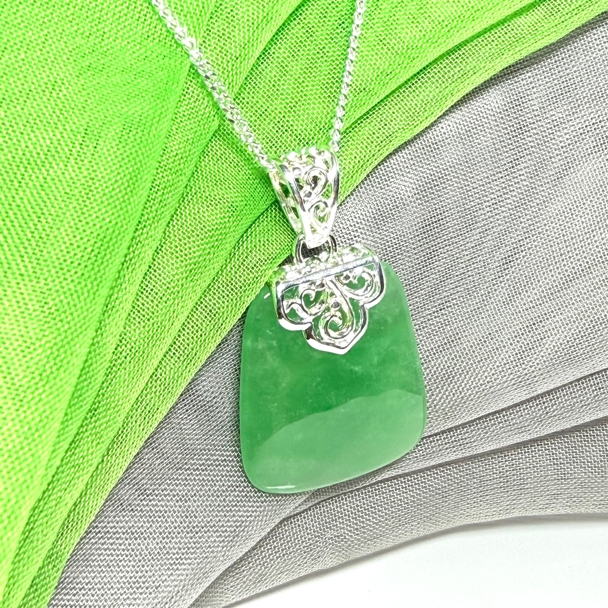 A green jadeite pendant necklace with a silver filigree bail, displayed on a textured green and gray fabric background.
